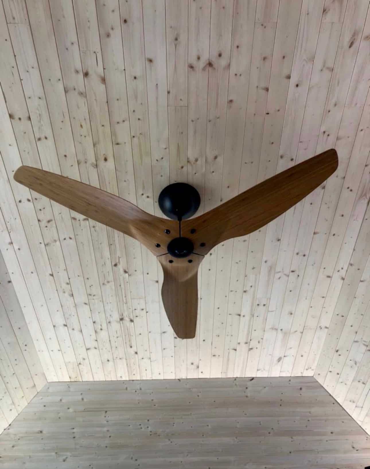 Three-blade wood ceiling fan with black motor housing installed on a plank ceiling